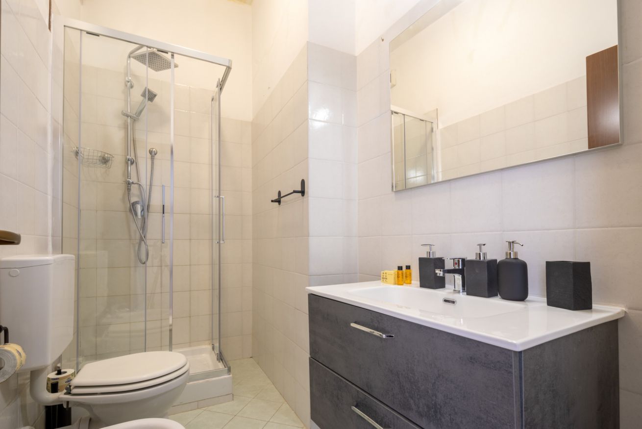 A modern bathroom with a glass-enclosed shower, a white toilet, and a dark-toned vanity with a mirror and toiletries.