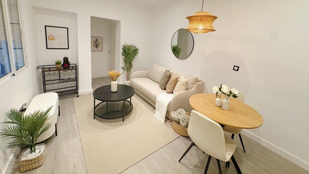 Ein modernes Wohnzimmer mit einer Couch, einem Couchtisch und einem Esstisch. Ein Fenster, eine Pflanze und ein Spiegel sind an der Wand zu sehen.