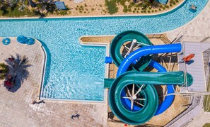 Vista aerea di una piscina con scivolo acquatico, circondata da un solarium piastrellato e palme.