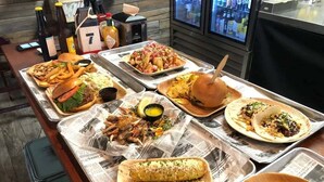 Una mesa de buffet con diversos platos, incluyendo hamburguesas, papas fritas, tacos y un elote.