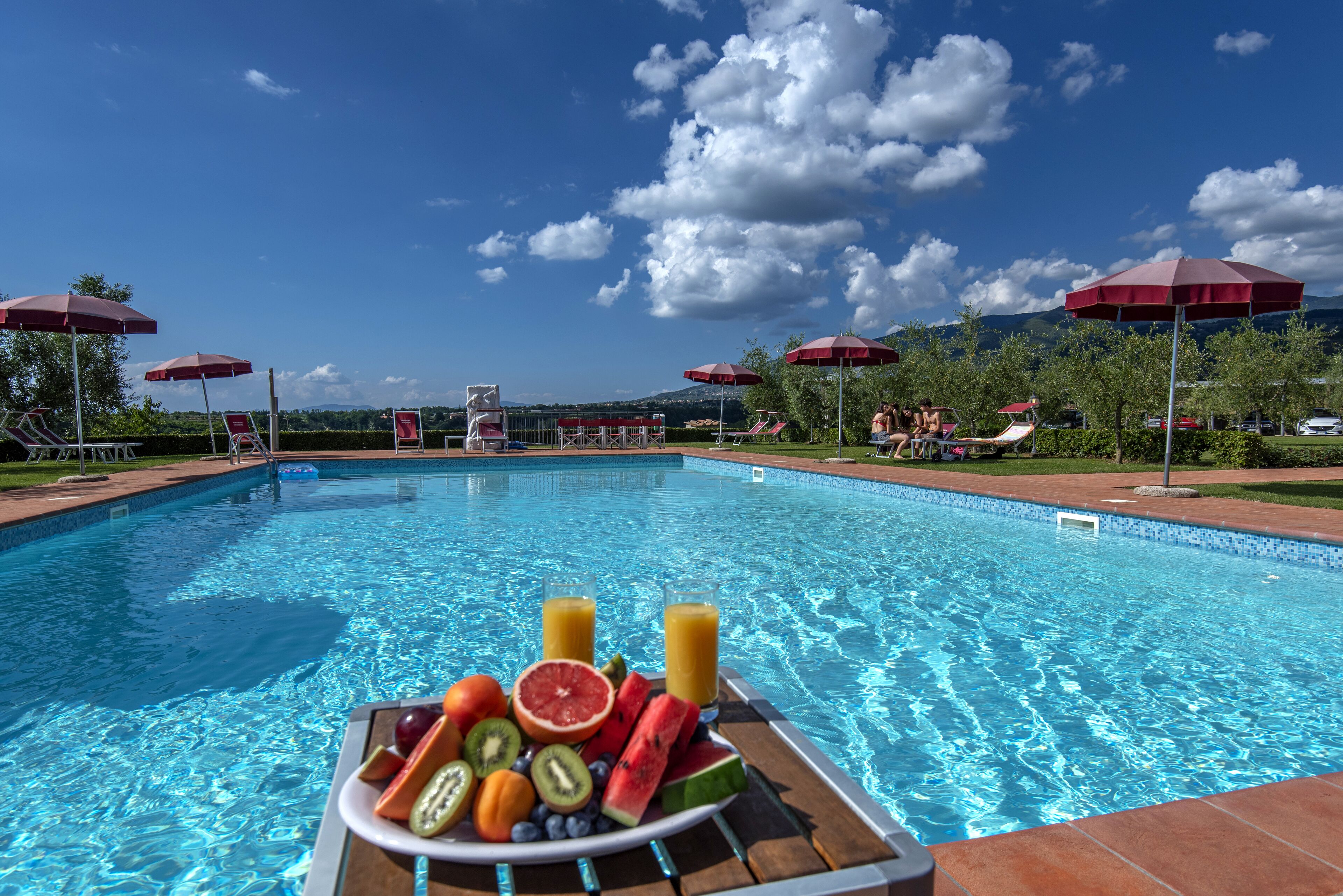 A swimming pool with a fruit and juice tray in the foreground, surrounded by lounge chairs and umbrellas.