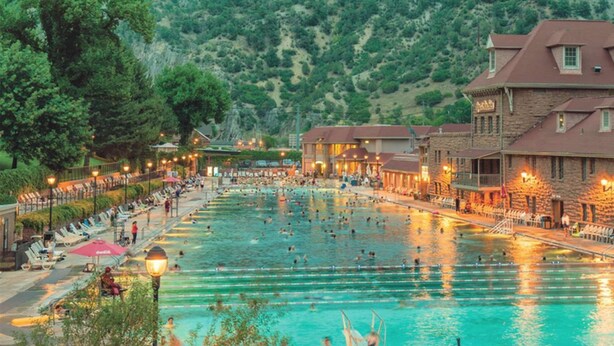 Una amplia piscina exterior con múltiples carriles, rodeada de sillones de descanso y sombrillas, con edificios y árboles al fondo.