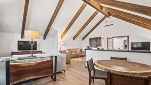 Un salón tipo ático con vigas de madera a la vista, una mesa de centro de cristal, un sofá, una mesa de comedor con sillas y un televisor.