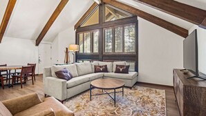 Una sala de estar estilo loft con techo alto, vigas de madera a la vista, un gran ventanal con contraventanas, un sofá modular con cojines decorativos, una mesa de centro redonda y un televisor sobre un mueble de madera.