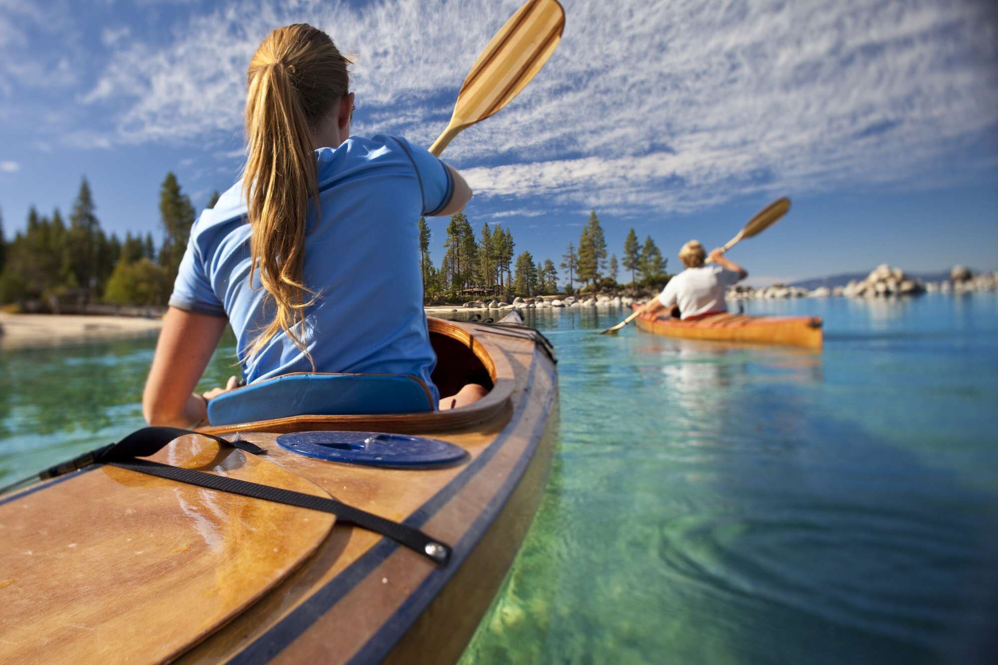 Two people kayaking on a calm body of water with clear skies and trees in the background.