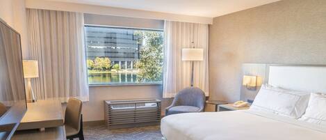 Una habitación de hotel con cama, escritorio, silla, calefacción y ventana con vista a un edificio y árboles.