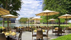 An outdoor patio area with wicker chairs and tables, large umbrellas, and a view of a building and a body of water in the background.