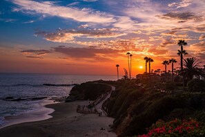 Vista de un atardecer costero con palmeras y un camino que lleva a un edificio.