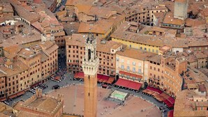 Vista aérea de uma praça histórica com uma torre imponente, rodeada por edifícios tradicionais com telhados de terracota.