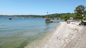A pebbled beach with boats in the water, a clear sky, and a distant town. - Vista Piazza by Interhome (San Felice del Benaco)