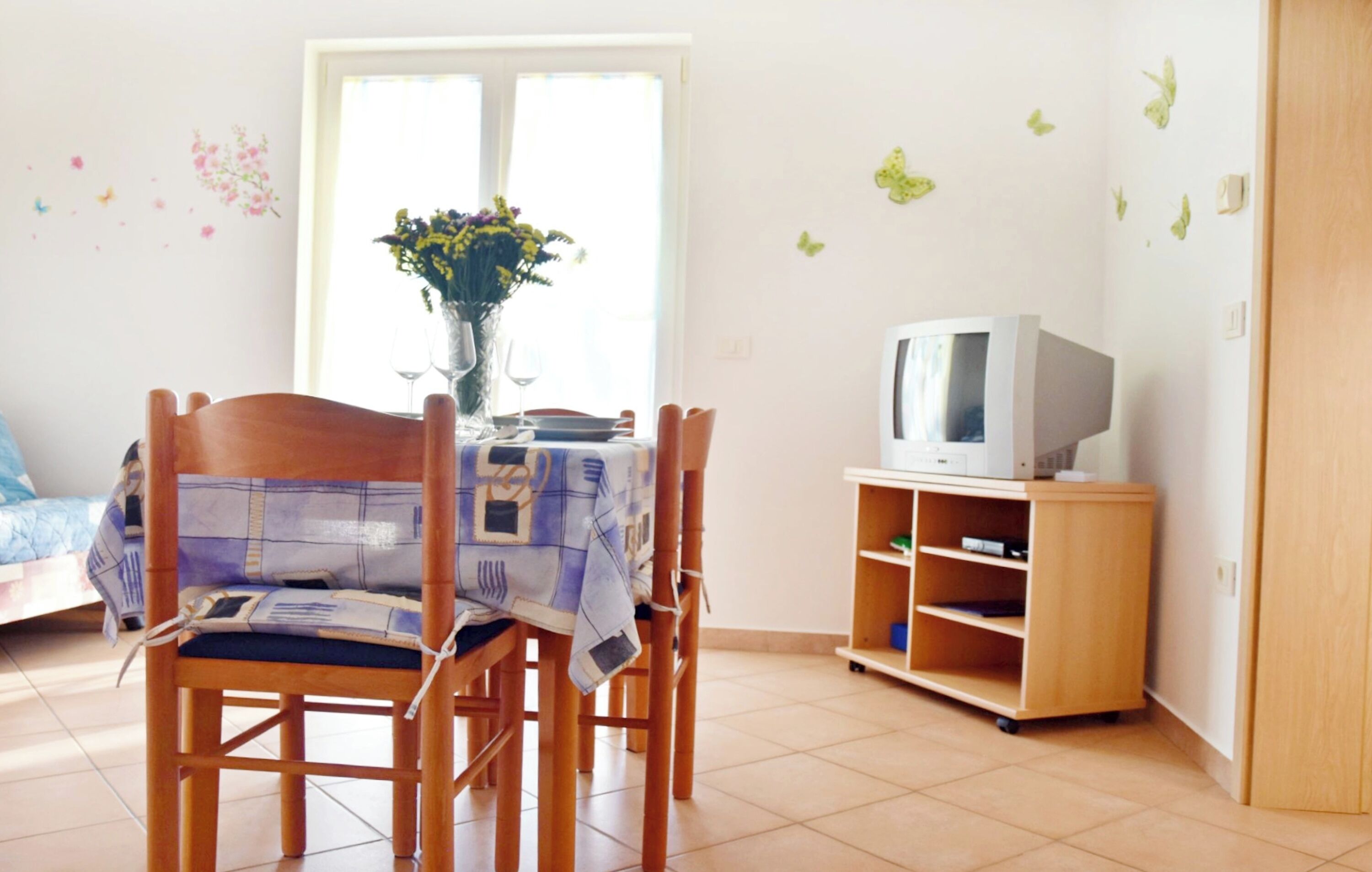 Ein Zimmer mit Esstisch für zwei Personen, einem Fernseher auf einem Holzständer und einem Fenster mit Blumen in einer Vase.