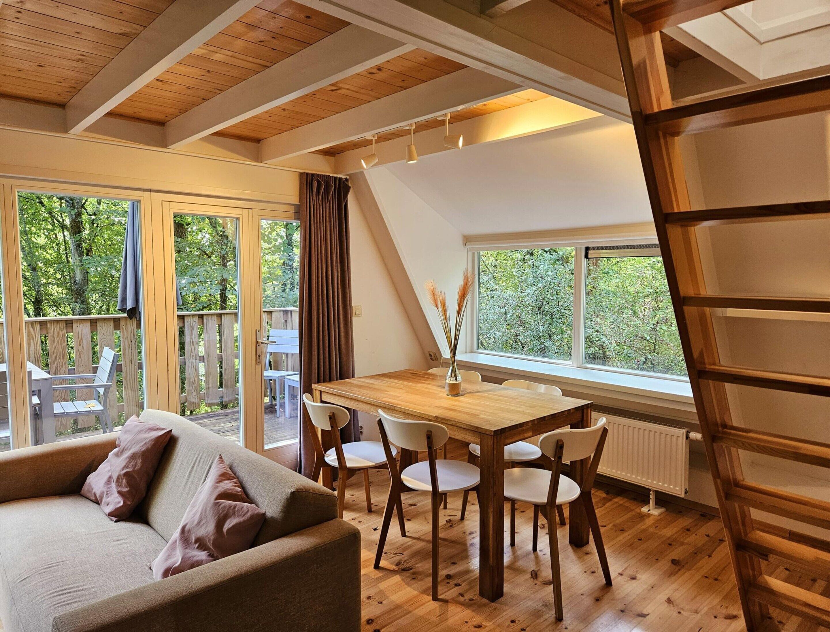 A cozy room with a wooden ceiling, a dining table with chairs, a sofa, and a view of the outdoors through large windows.