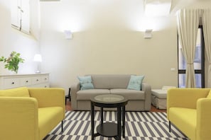 A living room with a sofa, two yellow armchairs, a glass-top coffee table, a striped rug, a white cabinet with a plant, and a window with curtains. - Santa Croce Deluxe Apartment (Firenze)