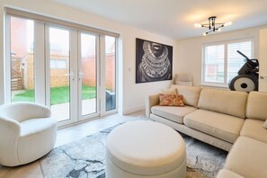 A spacious living room with a sliding glass door leading to a garden, a beige sofa, a white armchair, and a treadmill. - Host and Stay The Cosy Corner (Canterbury)
