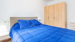 A bedroom with a bed, two pillows, a wooden wardrobe, and a nightstand with a lamp. - Apartment  by Interhome (St Antoni de Calonge)