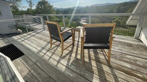 Una terraza de madera con dos sillas de mimbre y una mesa, con vistas a un paisaje pintoresco con montañas y un río.