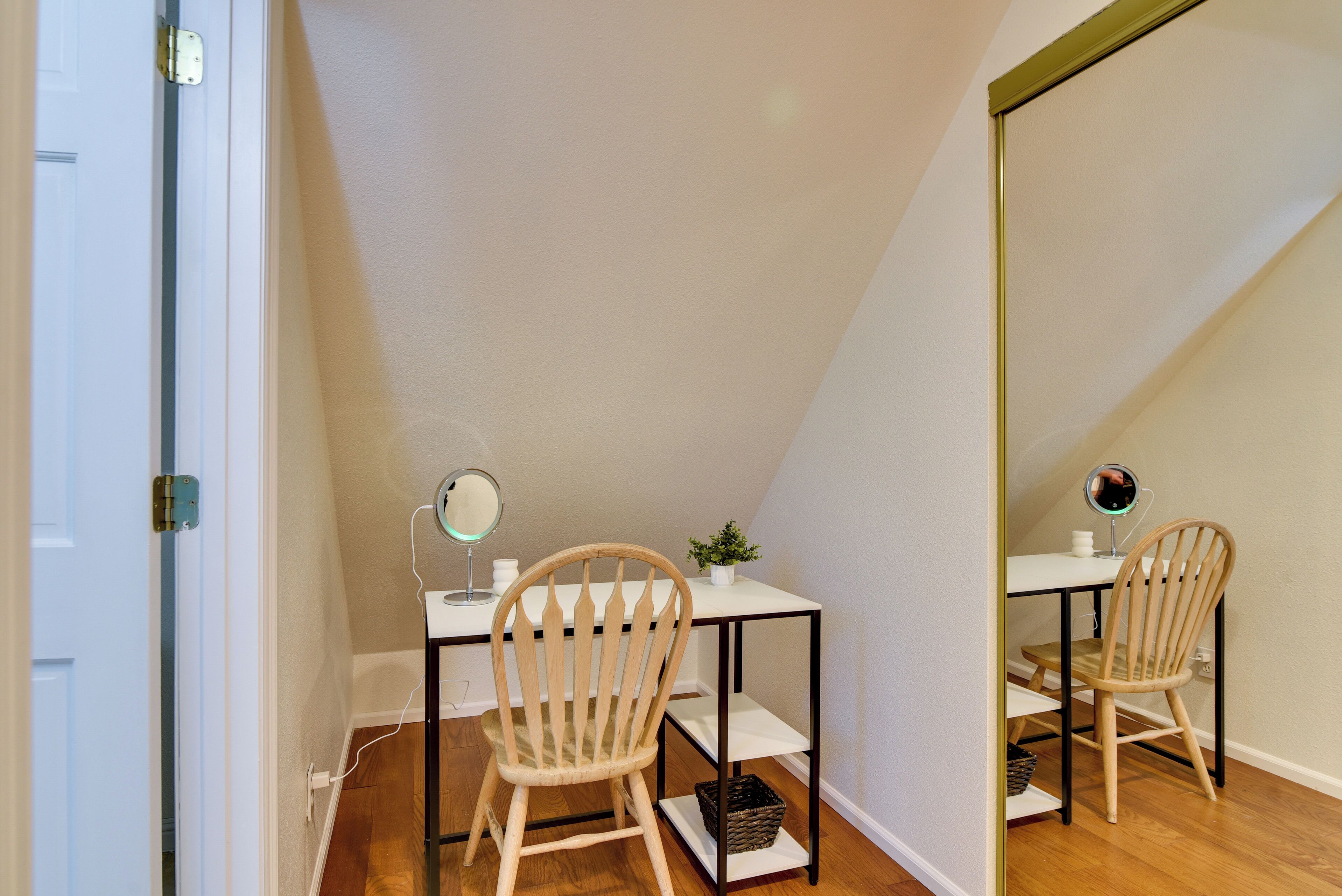 Una habitación pequeña y bien iluminada con escritorio, dos sillas, un espejo y una planta pequeña.
