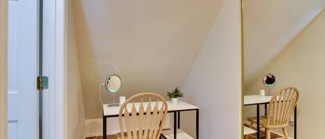 Una habitación pequeña y bien iluminada con escritorio, dos sillas, un espejo y una planta pequeña.
