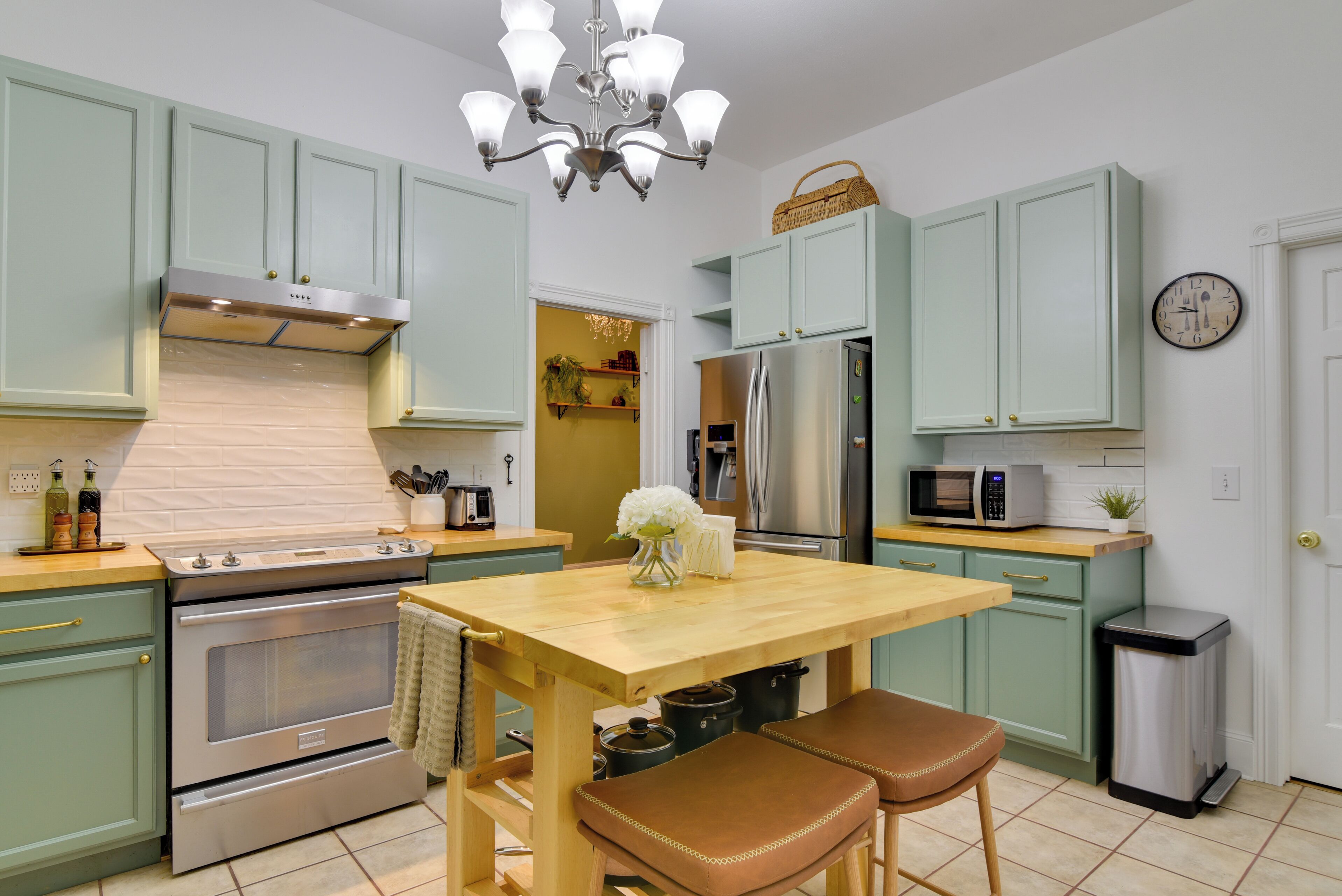 Une cuisine avec des meubles de couleur vert clair, des appareils en acier inoxydable, une table en bois et des chaises en cuir marron.