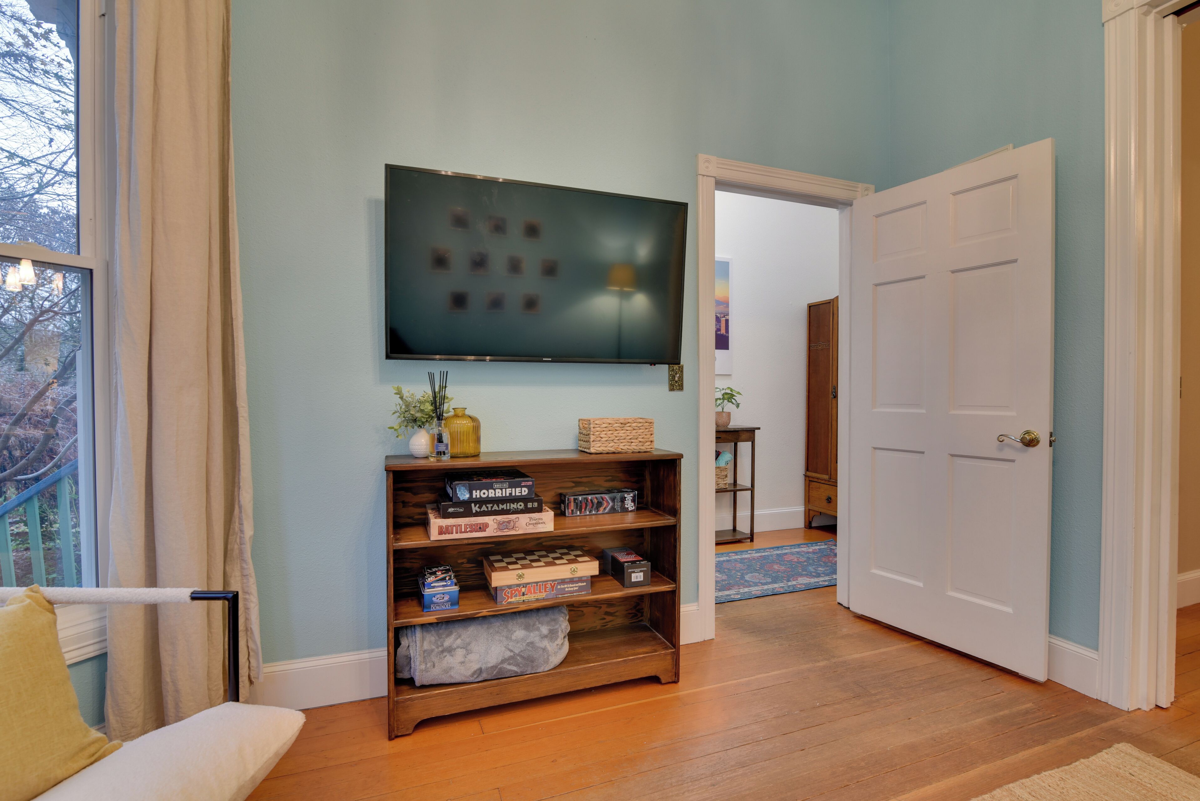 Een woonkamer met een houten vloer, een bank, een aan de muur bevestigde televisie en een boekenkast met diverse spullen.