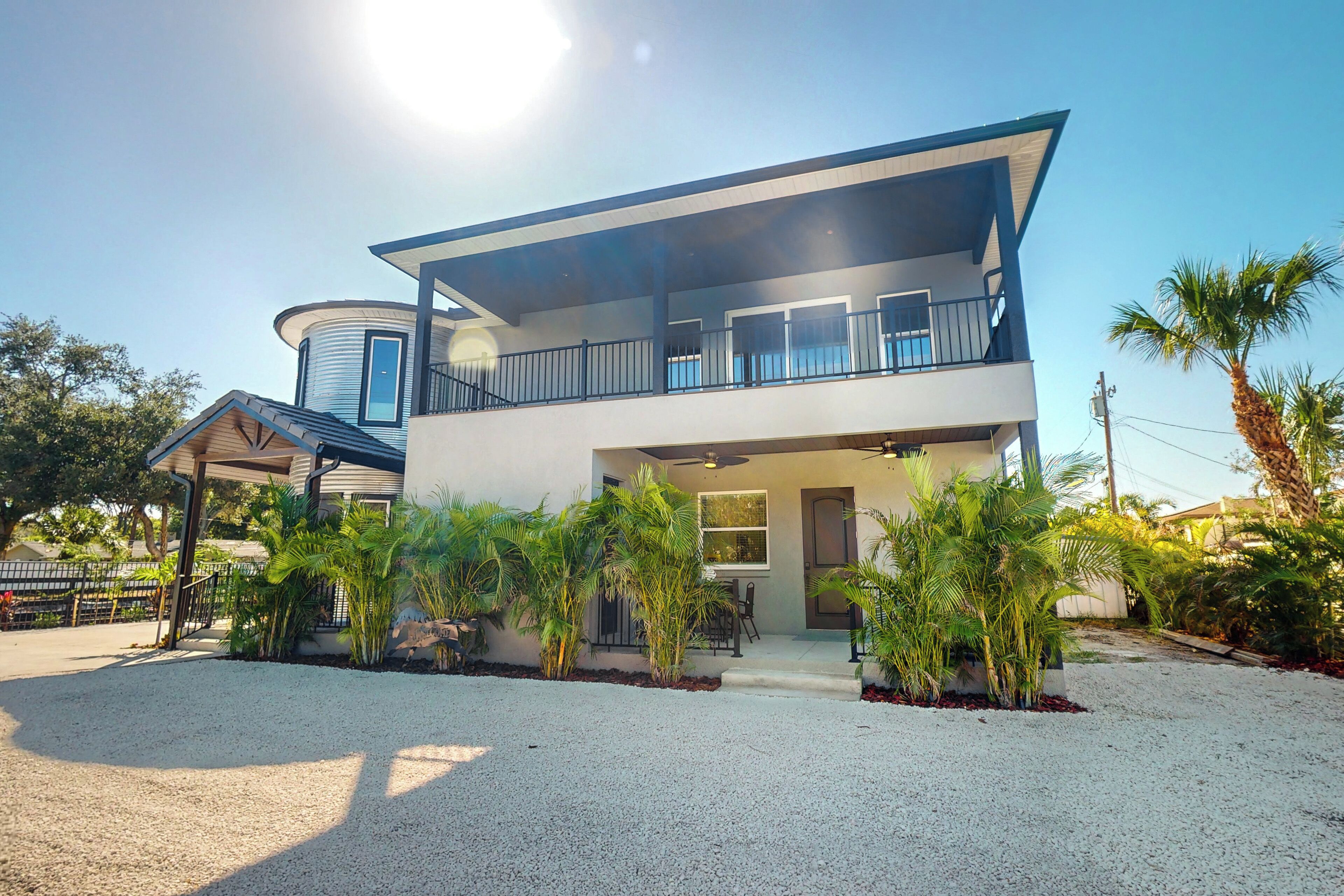 A modern two-story house with a balcony, surrounded by gravel and palm trees.