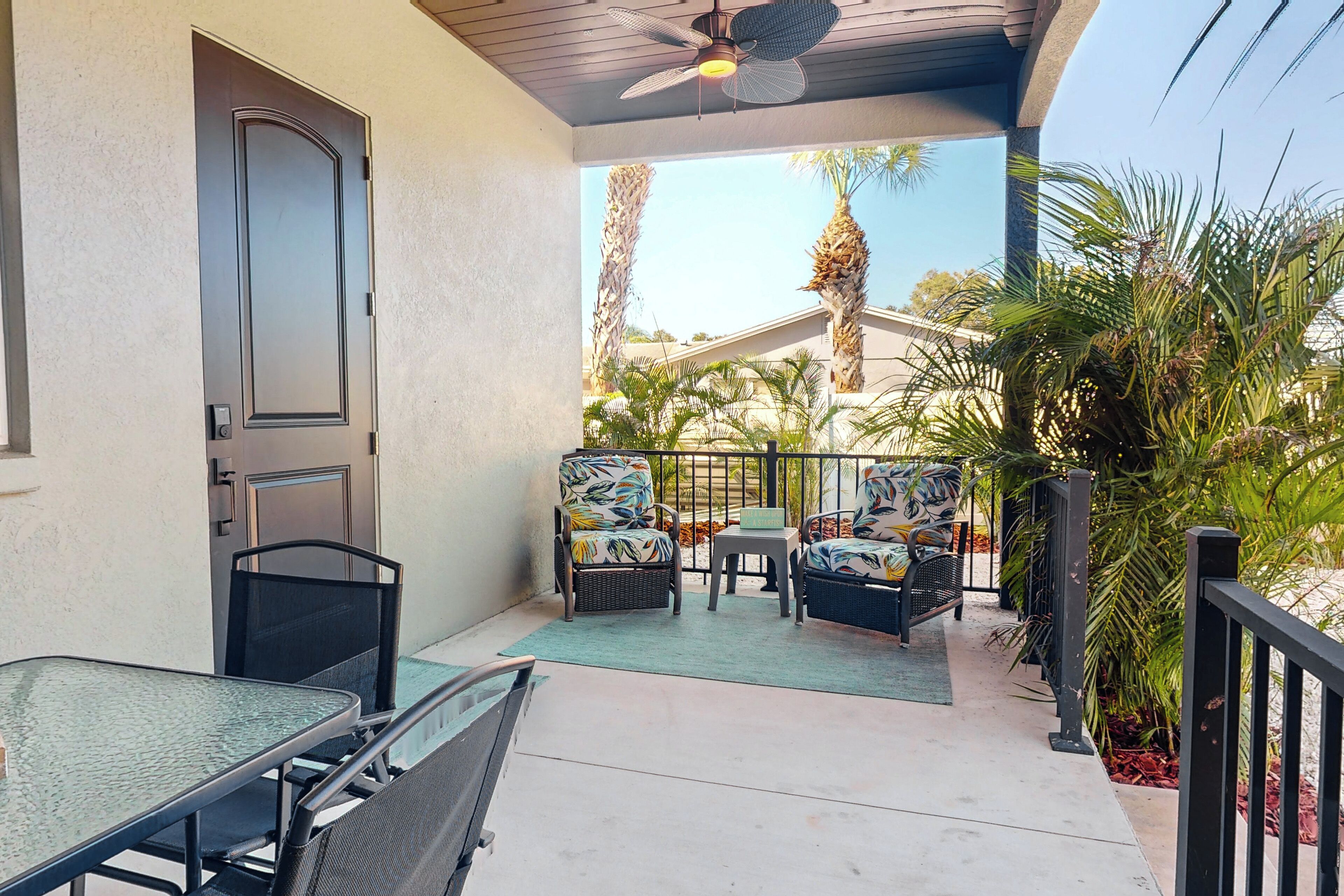 A covered patio with a table and chairs, a ceiling fan, and a view of palm trees and a house in the background.