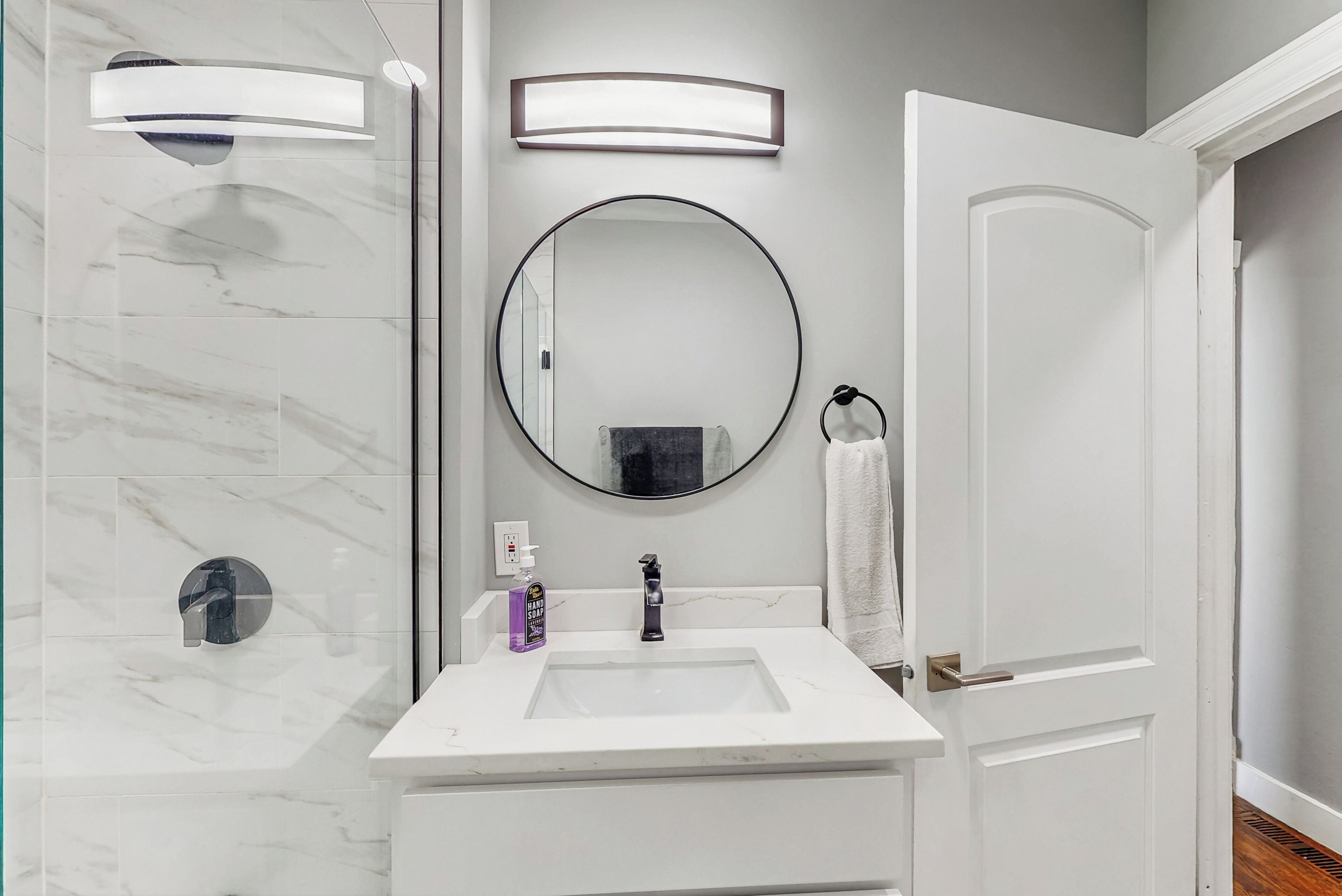 A bathroom with a white sink, a round mirror, a towel rack, and a glass shower enclosure.
