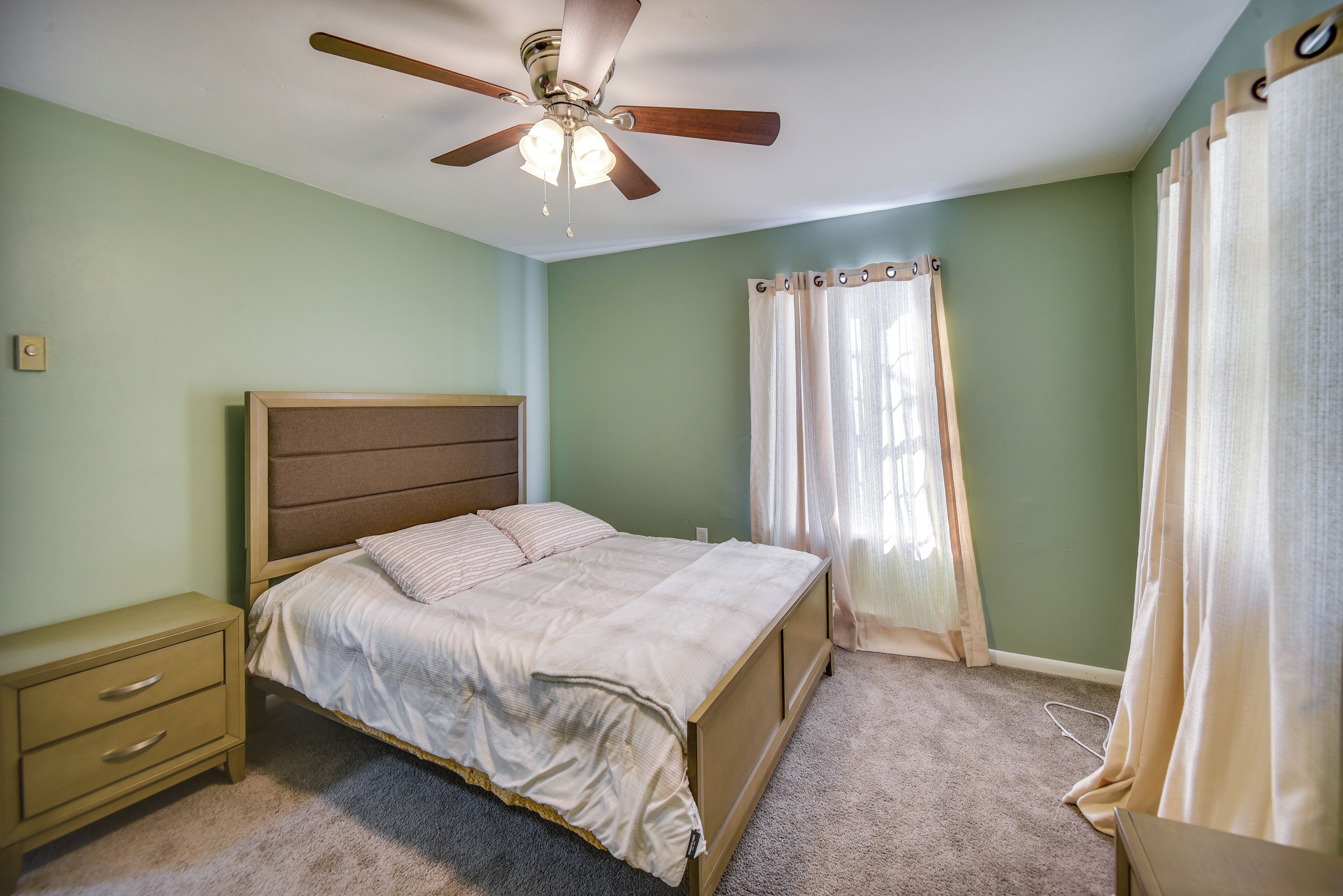 Un dormitorio con cama, mesita de noche, ventilador de techo y cortinas.