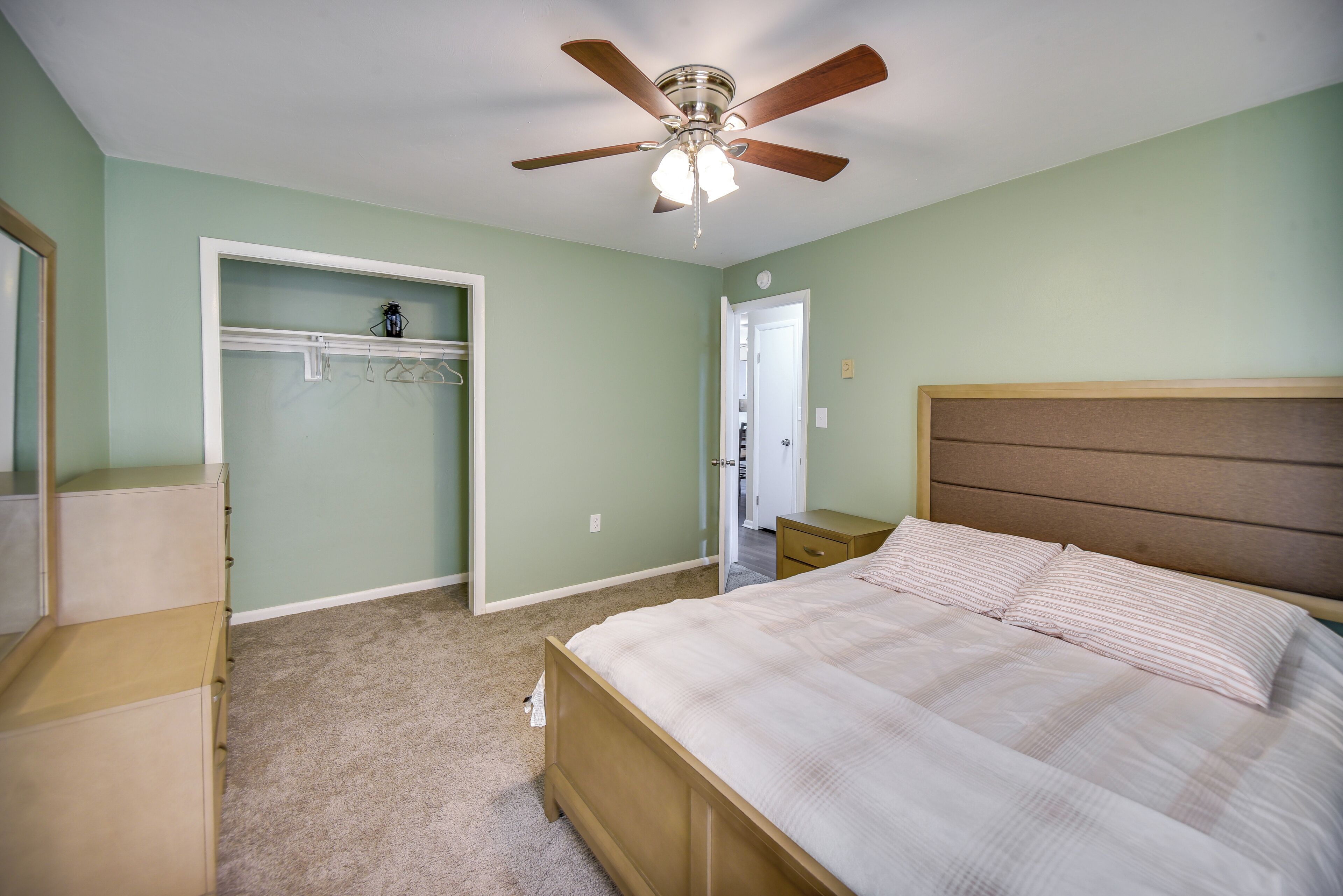 Une chambre à coucher comprenant un lit, un ventilateur de plafond, un dressing et une porte donnant sur une autre pièce.