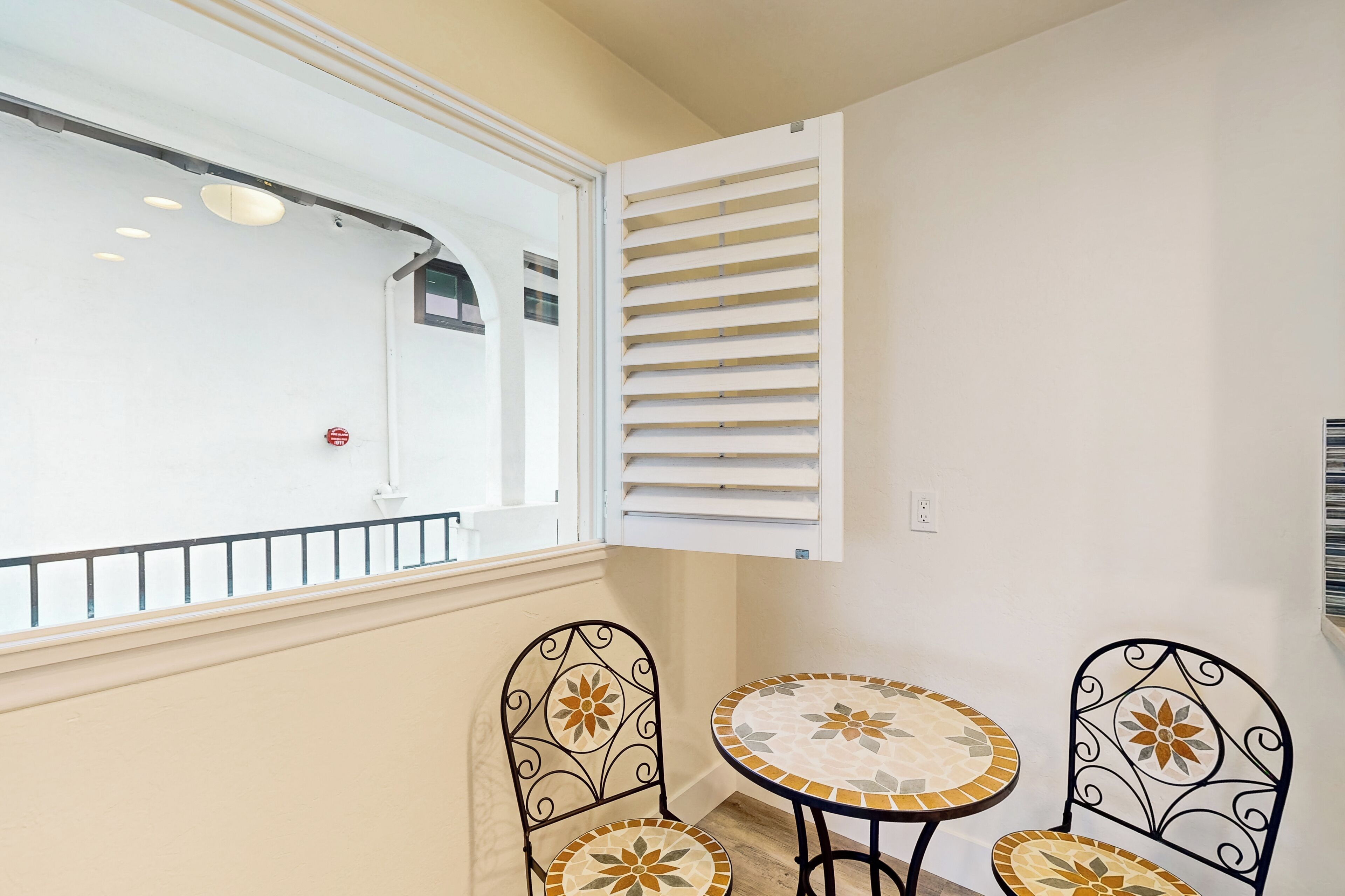 A balcony with two round tables and chairs, a white window with blinds, and a white wall.