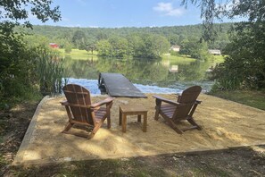 Een houten terras met twee stoelen en een tafel, met uitzicht op een meer, omringd door weelderige begroeiing en huizen in de verte.