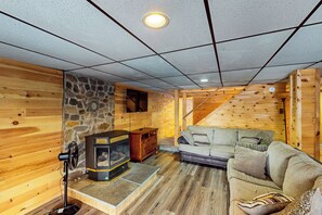 A cozy cabin interior with a stone fireplace, wooden walls, a sofa, and a staircase. - Spacious Lakefront Home w/ Private Hot Tub (White Haven)