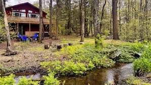 A cabin with a deck and outdoor seating, surrounded by a forest and a stream. - Spacious Lakefront Home w/ Private Hot Tub (White Haven)