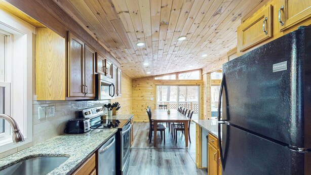 A kitchen with wooden cabinets, a black refrigerator, granite countertops, and a dining area with chairs and a table. - Spacious Lakefront Home w/ Private Hot Tub (White Haven)