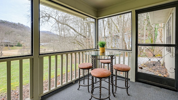 A covered outdoor seating area with a glass table and four stools, overlooking a landscaped garden and distant hills.
