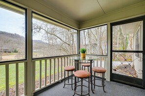 Un espace extérieur abrité, doté d’une table en verre et de quatre tabourets, offrant une vue sur un jardin aménagé et sur des collines lointaines.