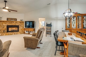 A living room with a stone fireplace, a television, a dining table with chairs, and a ceiling fan.