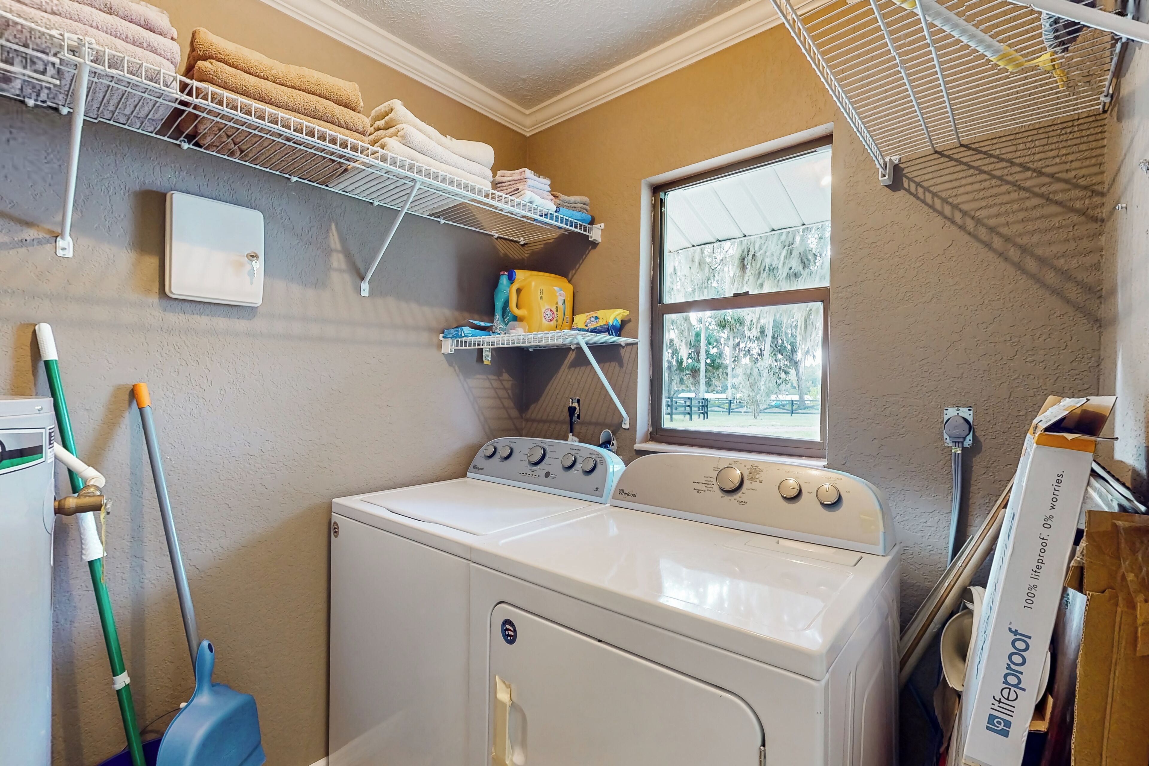 A laundry room with a front-loading washer and dryer, shelves with folded towels, a window, and a mop.