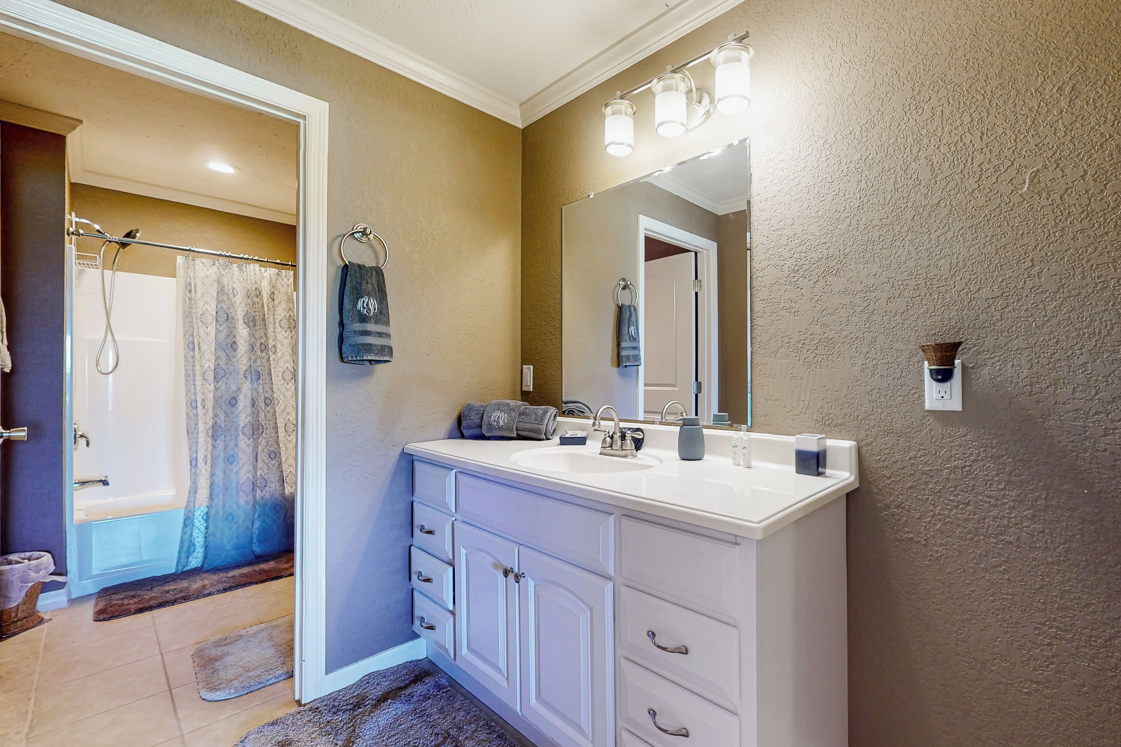 Une salle de bain avec un meuble-lavabo blanc, un grand miroir et un espace douche visible à travers une porte ouverte.