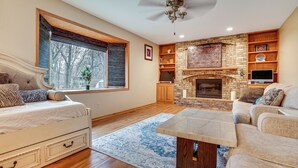 A spacious living room with a fireplace, a large window with blinds, a ceiling fan, and a television. - Rare Gem on a Ravine, Family Home in North Mankato (Mankato)