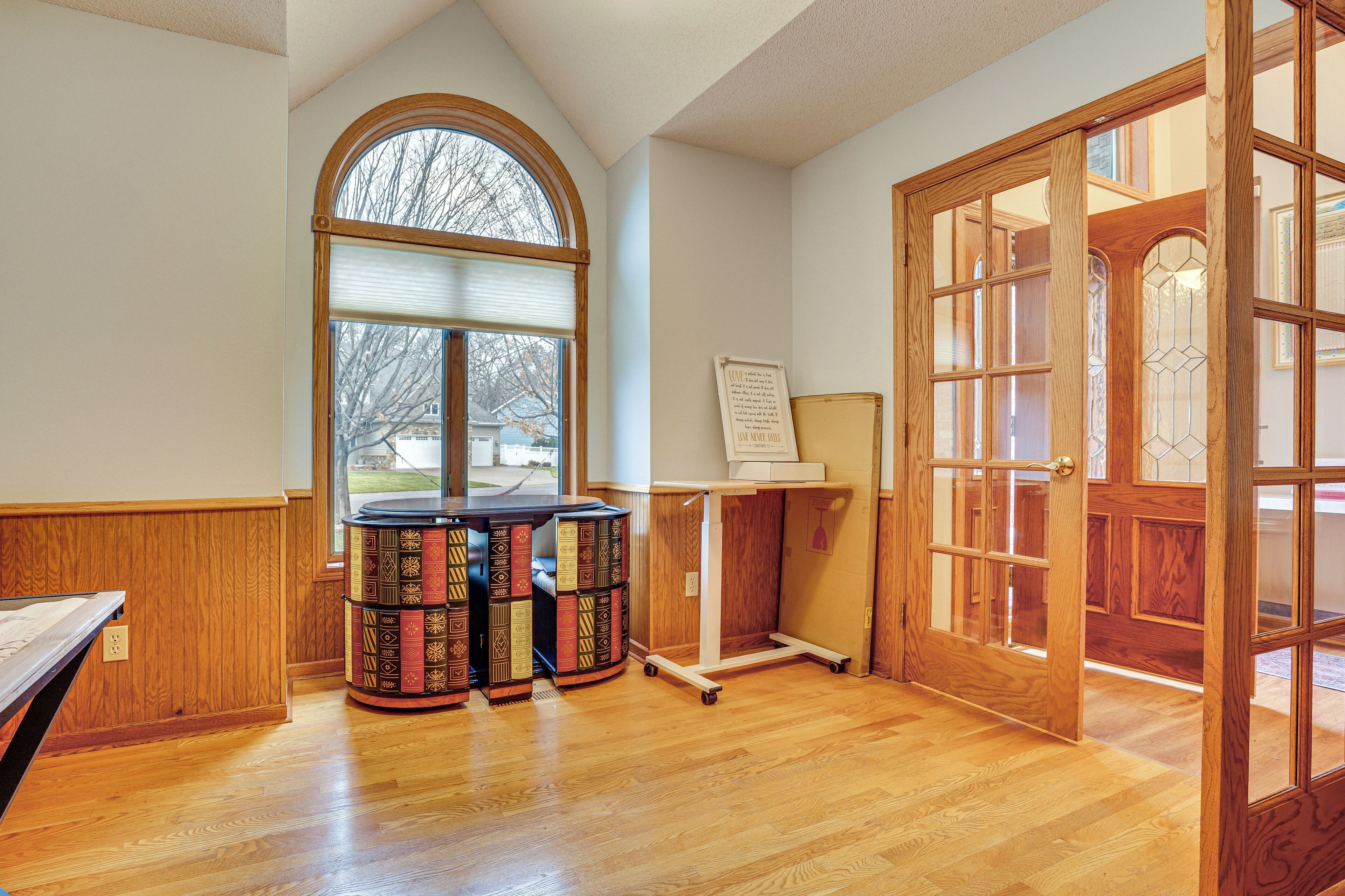 A room with a wooden floor, a large arched window, a wooden door with glass panels, and a small table with a bookcase.