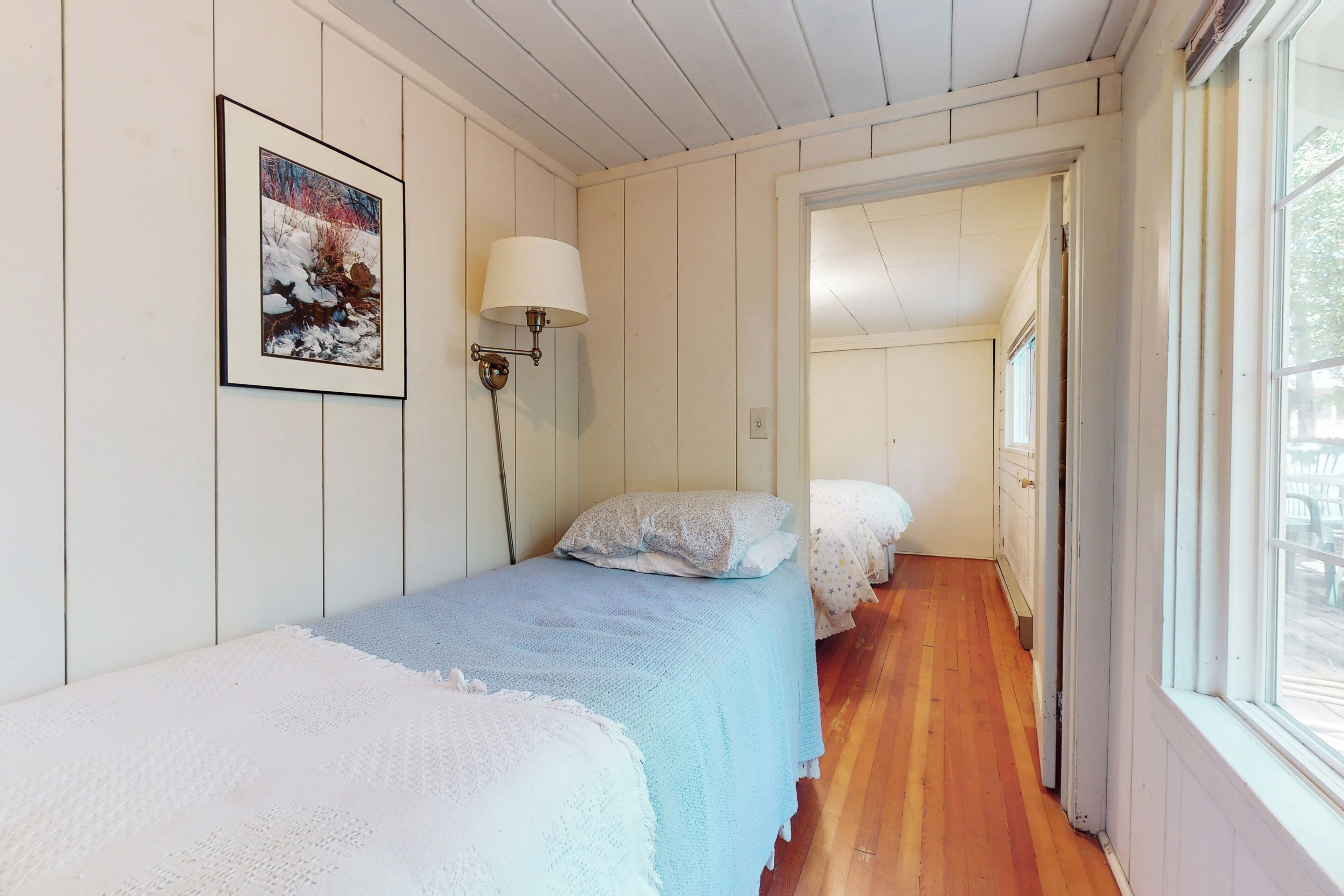 A narrow hallway with two beds, each with a white and blue blanket, and a framed picture on the wall.