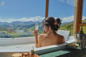 Une personne dans une baignoire, avec vue sur les montagnes, une bouteille et un bol de fraises.