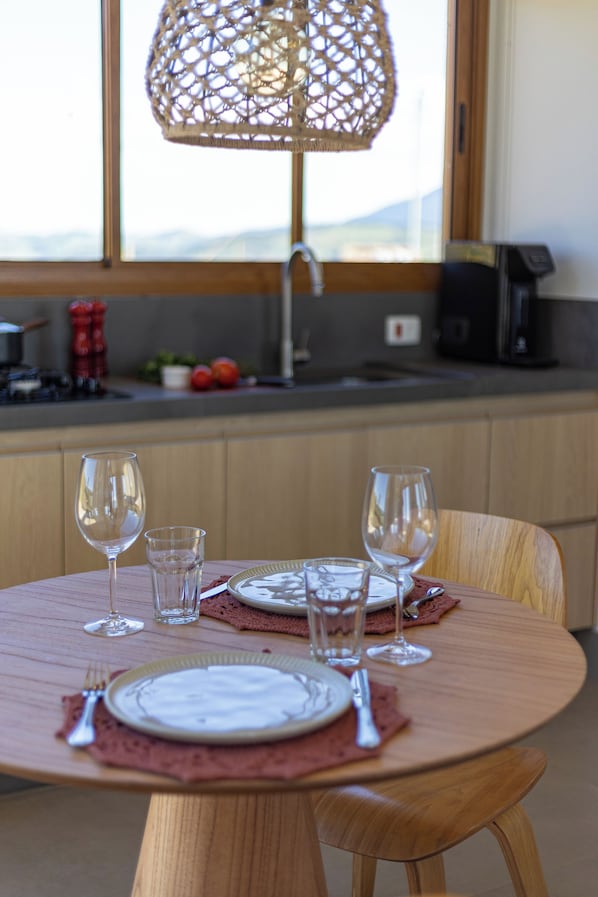 A dining table with plates, cutlery, and glasses set for two, a woven lamp, and a kitchen in the background. - Viso Villas Pousada Boutique (Sapucaí-Mirim)