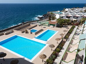 Un área de piscina en la azotea con dos piscinas, rodeada de sillones de descanso y sombrillas, con vista al océano.