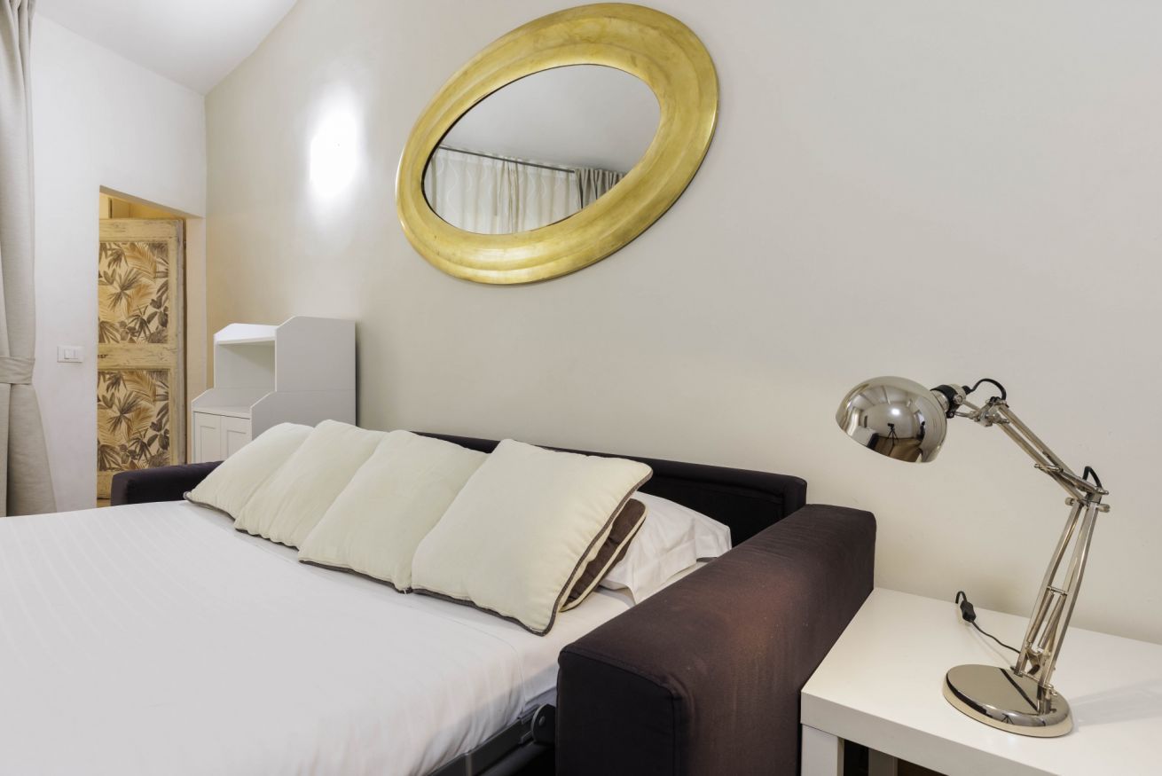 Une chambre d’hôtel avec un lit, une table de chevet avec une lampe, un miroir rond accroché au mur et une petite armoire blanche.
