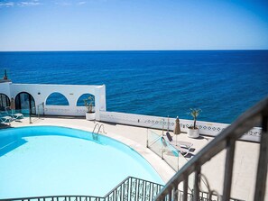 Ein Pool mit klarem, blauem Wasser, ein weißes Gebäude mit Rundbogenfenstern und Blick auf den Ozean.
