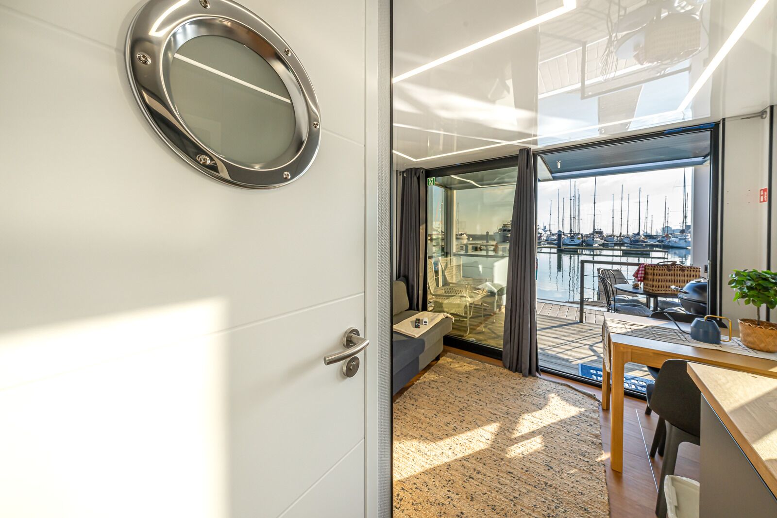 A modern interior with a large window overlooking a marina, a round mirror on the wall, and a small seating area with a table and chairs.
