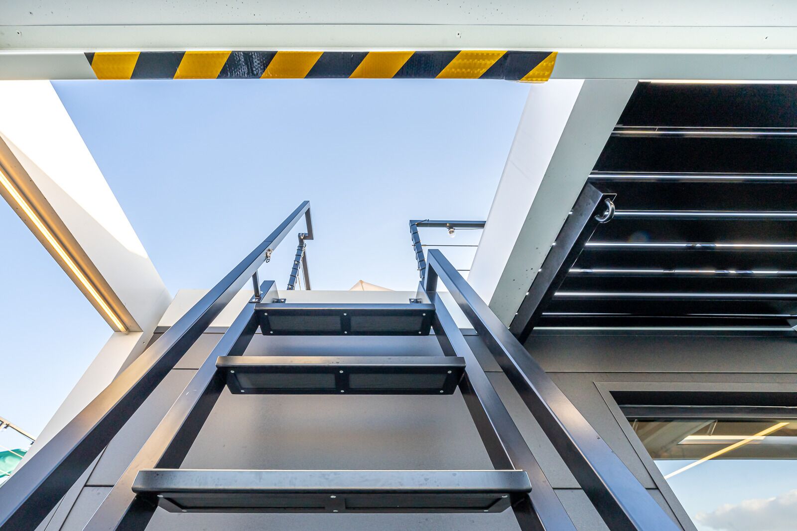 Un escalier métallique menant à un étage supérieur, avec une ligne de sécurité jaune et noire en bandes au sommet.
