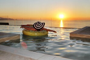 Eine Person in einem gelben aufblasbaren Schlauchboot, das in einem Pool bei Sonnenuntergang treibt. Im Hintergrund sind Ozean und Berge zu sehen.